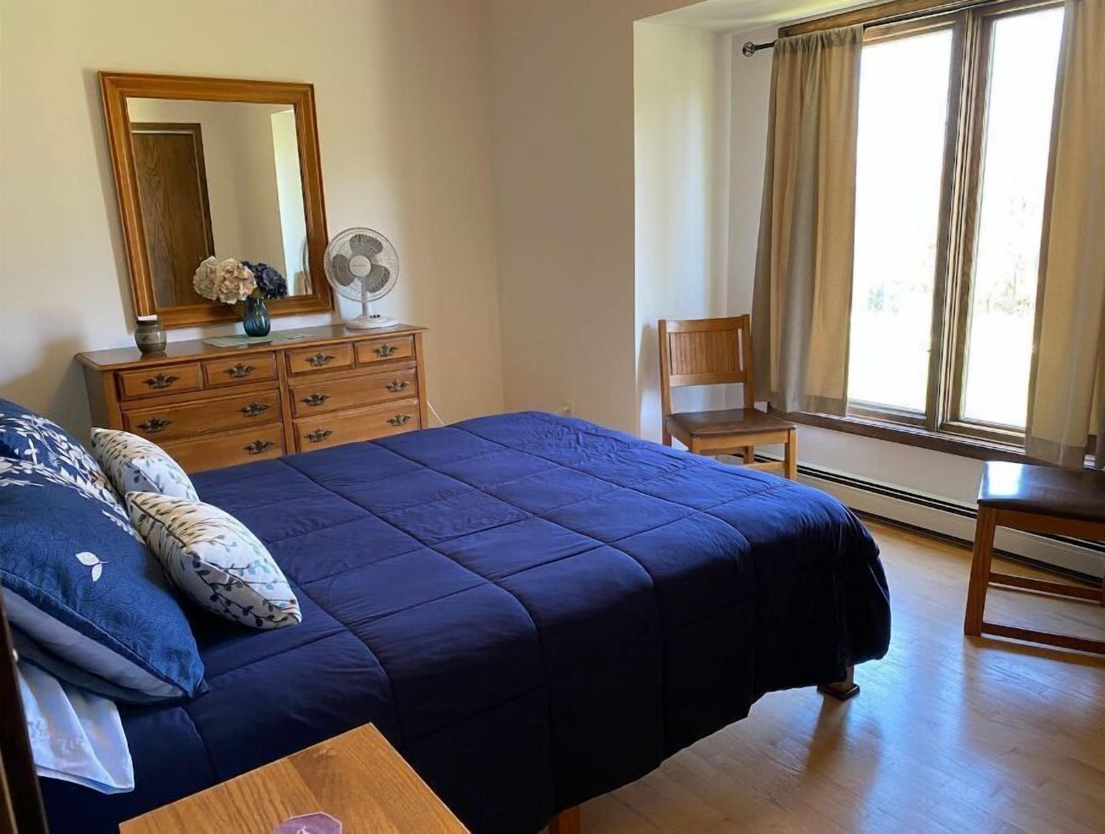 A bedroom with a large bed, a dresser, a chair, a window with curtains, and a fan.