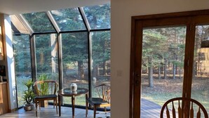 Ein Zimmer mit großen Fenstern, das einen Blick auf einen Wald bietet. Es gibt einen runden Tisch mit Stühlen, eine Topfpflanze und eine Vase auf dem Tisch.
