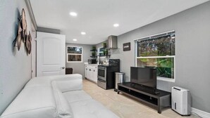 Una sala de estar moderna con un sofá blanco, un televisor de pantalla plana y una zona de cocina con microondas, horno y lavaplatos.