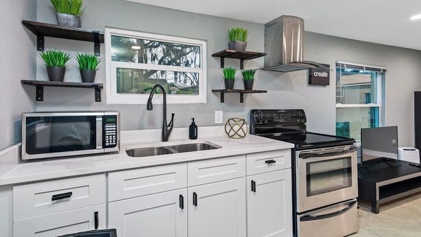 Una cocina moderna con armarios blancos, estufa de acero inoxidable, microondas y plantas en macetas sobre estantes.