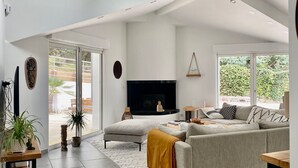 A modern living room with a grey sofa, a coffee table, and a flat-screen TV. There are two windows providing natural light and a view of greenery outside.