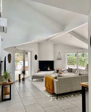 Un salon moderne avec un canapé gris, une table basse et un téléviseur à écran plat. Deux fenêtres laissent entrer la lumière naturelle et offrent une vue sur la verdure environnante.