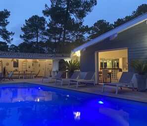 Zona de piscina con sillas de descanso, una piscina y un edificio con puerta abierta que da a un área de bar.