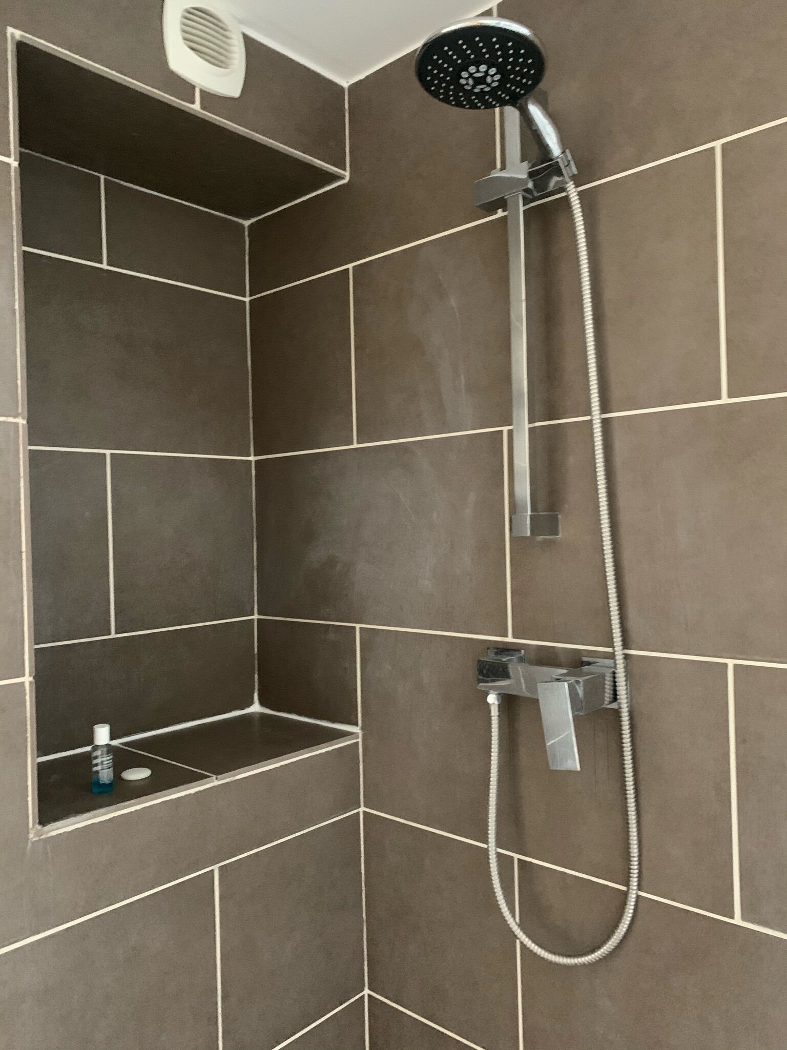 A shower with a handheld showerhead and a wall-mounted faucet, with a shelf below.