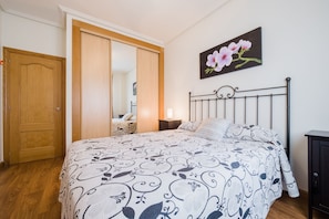 Een slaapkamer met een bed, een houten kledingkast, een nachtkastje, een lamp en een schilderij van bloemen aan de muur.
