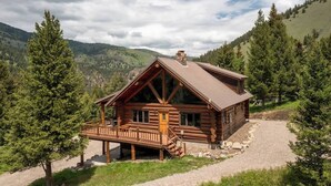 A log cabin with a porch and a chimney, surrounded by trees and a hilly landscape.