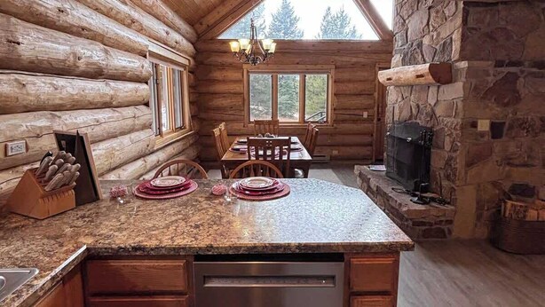 A log cabin interior with a kitchen area featuring a granite countertop, wooden cabinets, a built-in dishwasher, and a stone fireplace.