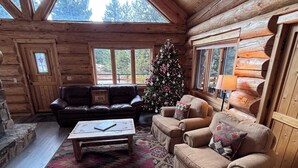 A log cabin interior with a Christmas tree, leather sofas, a coffee table, and a patterned rug.