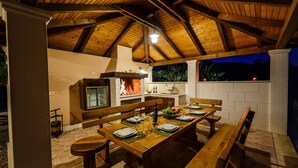 Un área de comedor al aire libre con una mesa de madera dispuesta para seis personas, una parrilla y un muro de piedra como fondo.