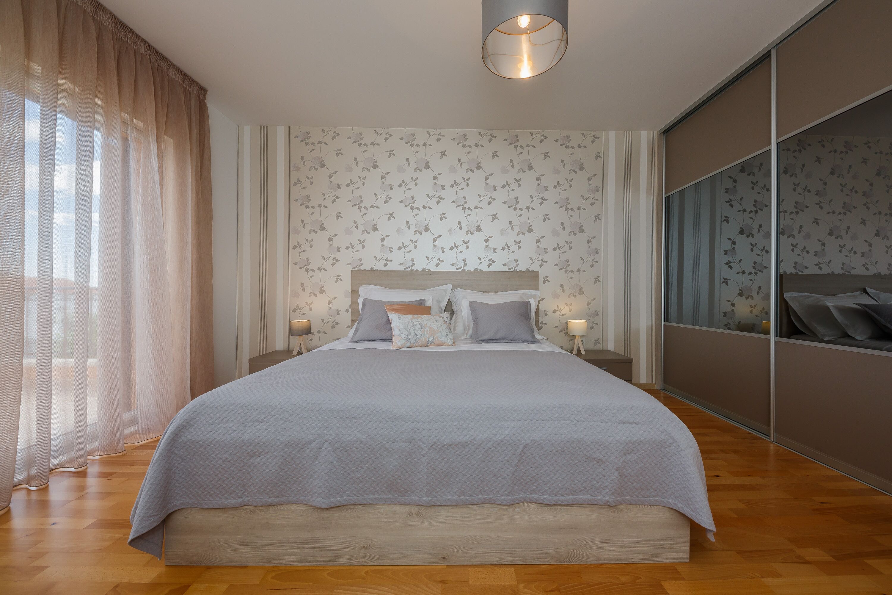 Een moderne slaapkamer met een groot bed, houten vloeren en een groot raam met dunne gordijnen.