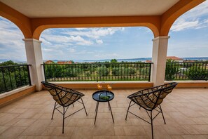 Ein Balkon mit zwei Korbstühlen und einem kleinen Tisch, der auf eine grüne Landschaft mit weit entfernten Gebäuden blickt.