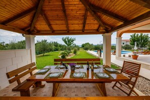 Eine Terrasse im Freien mit einem Holztisch, gedecktem Esstisch, Bänken und Blick auf einen angelegten Garten und einen Poolbereich.