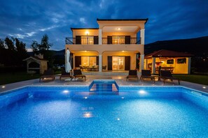 A two-story house with a swimming pool in front, featuring outdoor seating and a patio area.