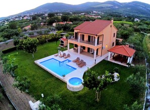 Ein zweistöckiges Haus mit Swimmingpool, einer Terrasse mit Liegestühlen und Blick auf die umliegende Landschaft.