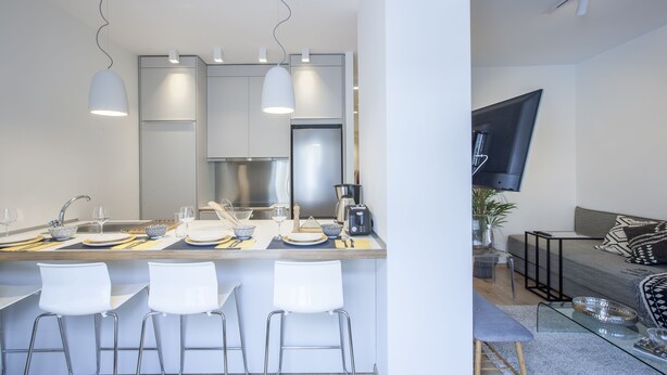 A modern kitchen with bar stools, a dining table set for four, and a living area with a sofa and TV. - Alterra Vita City Apartment (Thessaloniki)