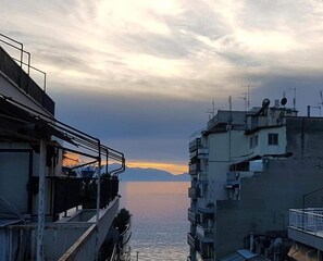 A coastal cityscape with buildings overlooking a calm sea and distant mountains. - Alterra Vita City Apartment (Thessaloniki)