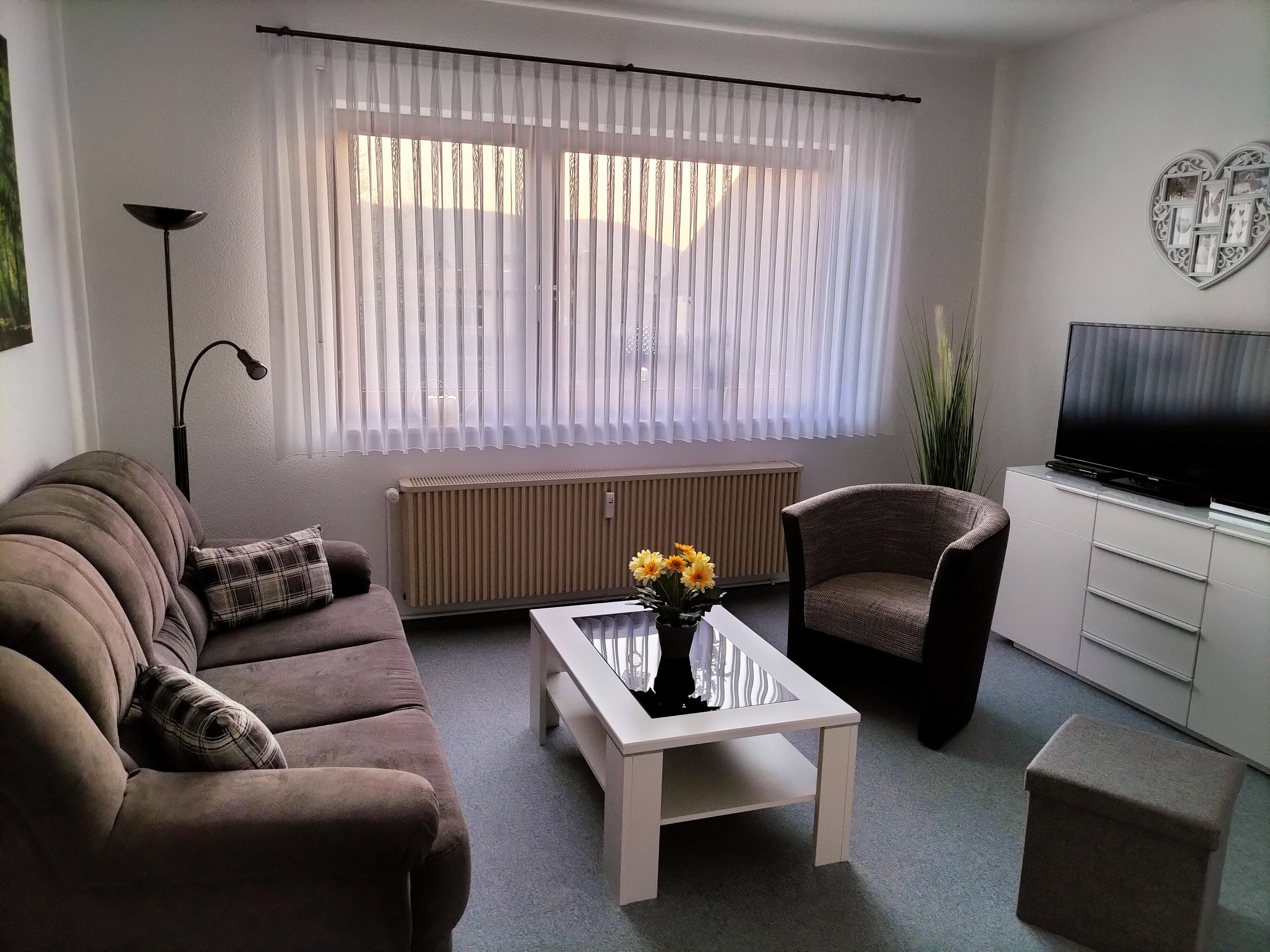 A living room with a brown sofa, a white coffee table with flowers, a brown armchair, a white cabinet with a TV, and a radiator.