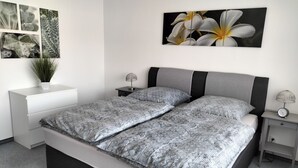 Une chambre moderne avec une couvre-lit à motifs gris et blancs, une table de chevet blanche ornée d’une plante, une tête de lit grise et une œuvre d’art représentant des fleurs blanches, accrochée au mur.