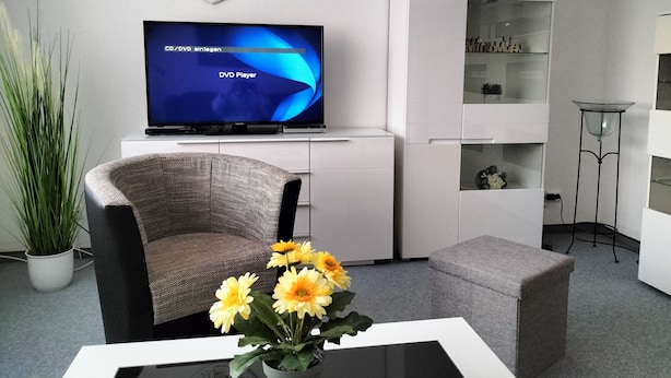 Un salon moderne avec un téléviseur à écran plat, un fauteuil gris, une table basse ornée d’un vase de fleurs jaunes et un meuble blanc.