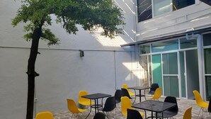 An outdoor patio area with tables and chairs, a tree, and a building with large windows. - El Gran Hostal (Guadalajara)