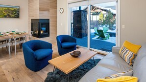 Una sala de estar moderna con chimenea, dos sillones azules, una mesa de centro de madera y un sofá gris con cojines amarillos.