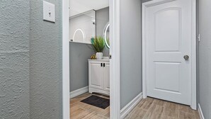 Un pasillo con una puerta blanca, un armario pequeño con un espejo y una planta en el mostrador.