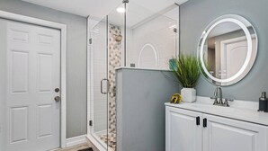 Un baño con puerta blanca, cabina de ducha de vidrio, tocador blanco con espejo y una planta en maceta.