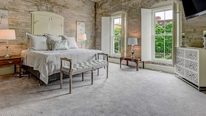A bedroom with a brick wall, a bed with a headboard, two bedside tables with lamps, a bench, and a TV mounted on the wall.