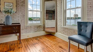 A room with exposed brick walls, large windows with views of a river and buildings, a wooden floor, a dark blue chair, and a dresser with a vase of flowers.