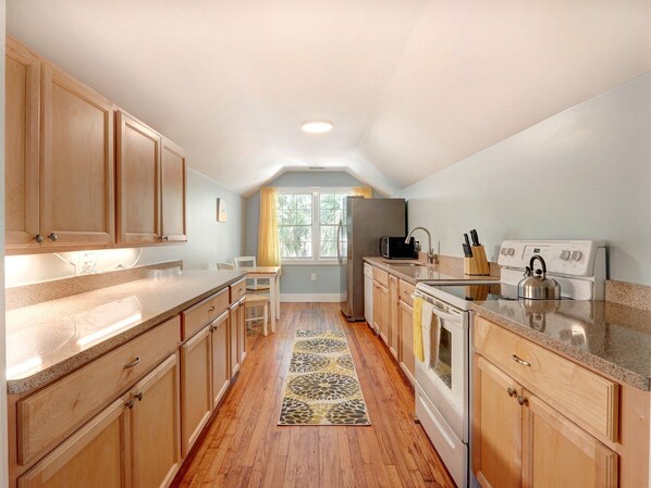 A kitchen with wooden cabinets, a granite countertop, a refrigerator, a stove, and a window with a view of greenery.