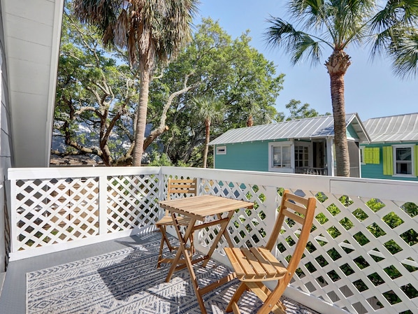 A balcony with a wooden table and chairs, a white lattice fence, and a view of a teal house with a green window. - Heated Pool Access, Large Tybee Home (Tybee Island)