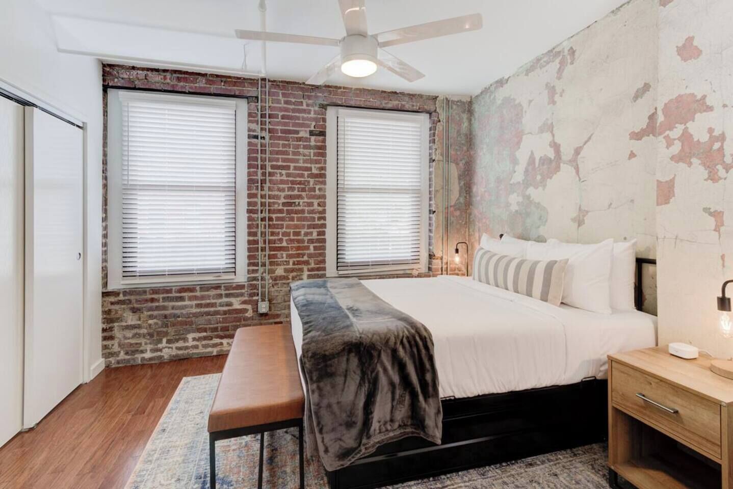 A bedroom with a brick wall, two windows with blinds, a bed with a white duvet, a bench, a bedside table with a lamp, and a ceiling fan.