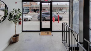 Entrada de un hotel con puerta de vidrio, una planta en maceta, un extintor y felpudo.