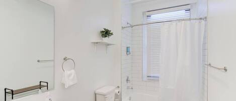 A modern bathroom with a white sink, toilet, and bathtub. There is a window with a shower curtain, a towel rack, and a small shelf with a plant.