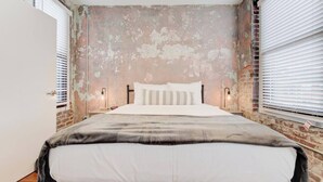 Un dormitorio con una cama, dos lámparas de noche, una pared de ladrillo y una ventana con persianas.