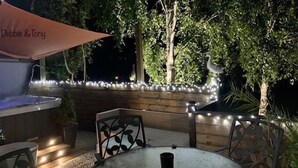 Un espace extérieur aménagé en terrasse, avec une table et des chaises, entouré d’arbres et bordé d’un mur en bois décoré de guirlandes lumineuses.
