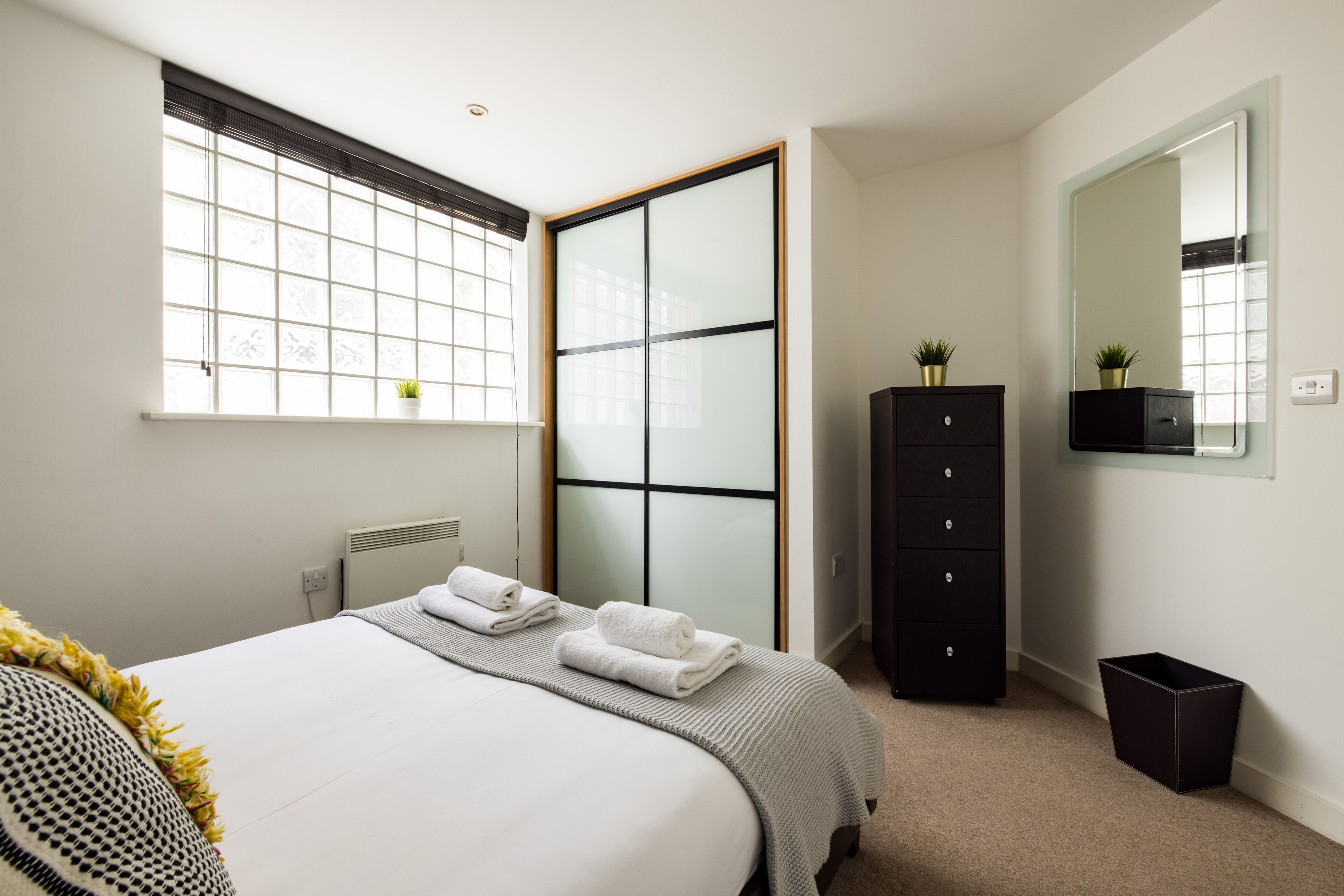 A modern bedroom with a large bed, a wardrobe, a mirror, and a window with blinds.