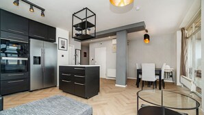 A modern kitchen with stainless steel appliances, a dining area with a glass table, and a living space with a gray sofa.