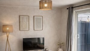 A modern living room with a flat-screen TV on a white console, a brown leather sofa, a large window with curtains, and a tall, woven pendant light. - King Size bed - Free Parking - 5 min to Centre (Lincoln)