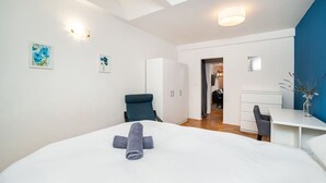 Une chambre d’hôtel moderne avec un grand lit, un bureau et une chaise. Deux tableaux ornent le mur et une lucarne se trouve au-dessus du lit.