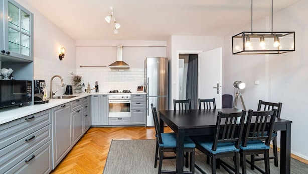 Une cuisine moderne avec des meubles gris, un réfrigérateur en acier inoxydable et un coin repas comprenant une table et des chaises en bois foncé.