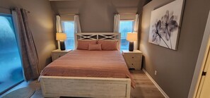 A bedroom with a bed, two bedside tables with lamps, a window with blinds, and a wall with a framed picture.