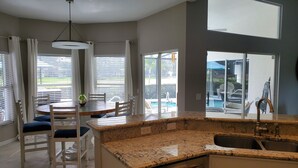 Eine Küche mit Granitarbeitsplatte, ein Essbereich mit Tisch und Stühlen sowie ein Blick auf einen Swimmingpool durch große Fenster.