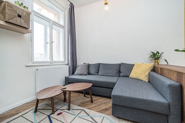 A modern living room with a grey sectional sofa, a small wooden table, a patterned rug, a radiator, and a window with curtains.