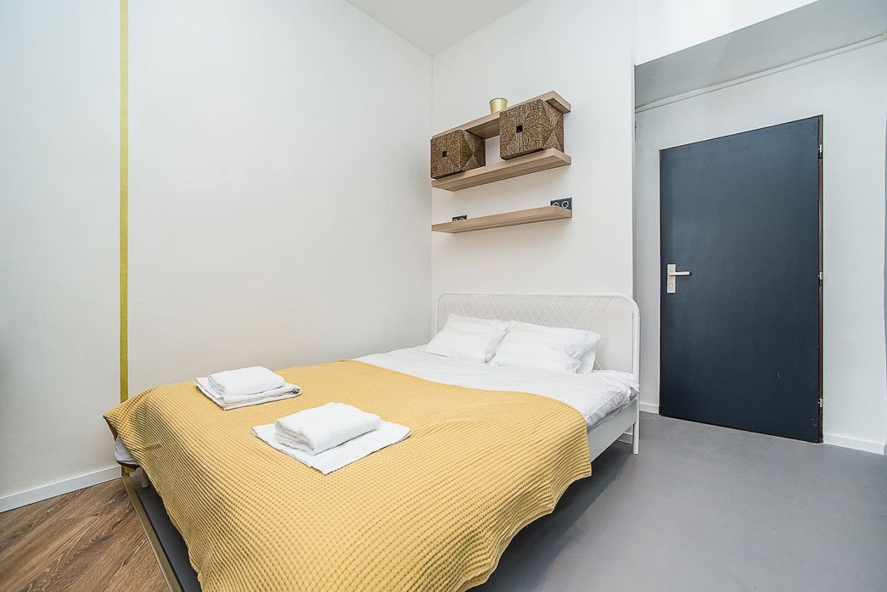 A single bed with a yellow blanket, white pillows, and a white headboard. There are two wooden shelves above the bed, and a dark door to the right.