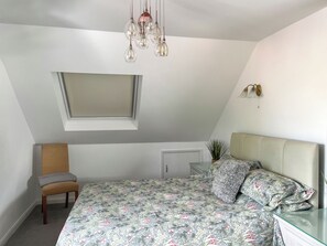 A bedroom with a sloped ceiling, a bed with floral bedding, a chair, a small window, and a wall-mounted light fixture.