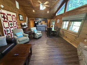 A cozy cabin interior with a wooden floor, a dining area, a kitchen, and a large window providing natural light.