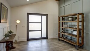Una stanza con pavimento in legno, una porta con pannelli trasparenti satinati, una libreria in legno con la scritta “HOME” e un tavolino con una lampada e una pianta.