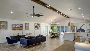 Un ampio soggiorno con moderna ventola a soffitto, una zona pranzo con tavolo e sedie in legno e una cucina con mobili bianchi ed elettrodomestici in acciaio inossidabile.