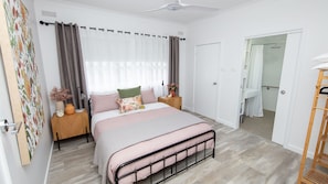 A bedroom with a bed, nightstands, a wooden floor, a ceiling fan, a bathroom with a shower, and a door leading to another room.